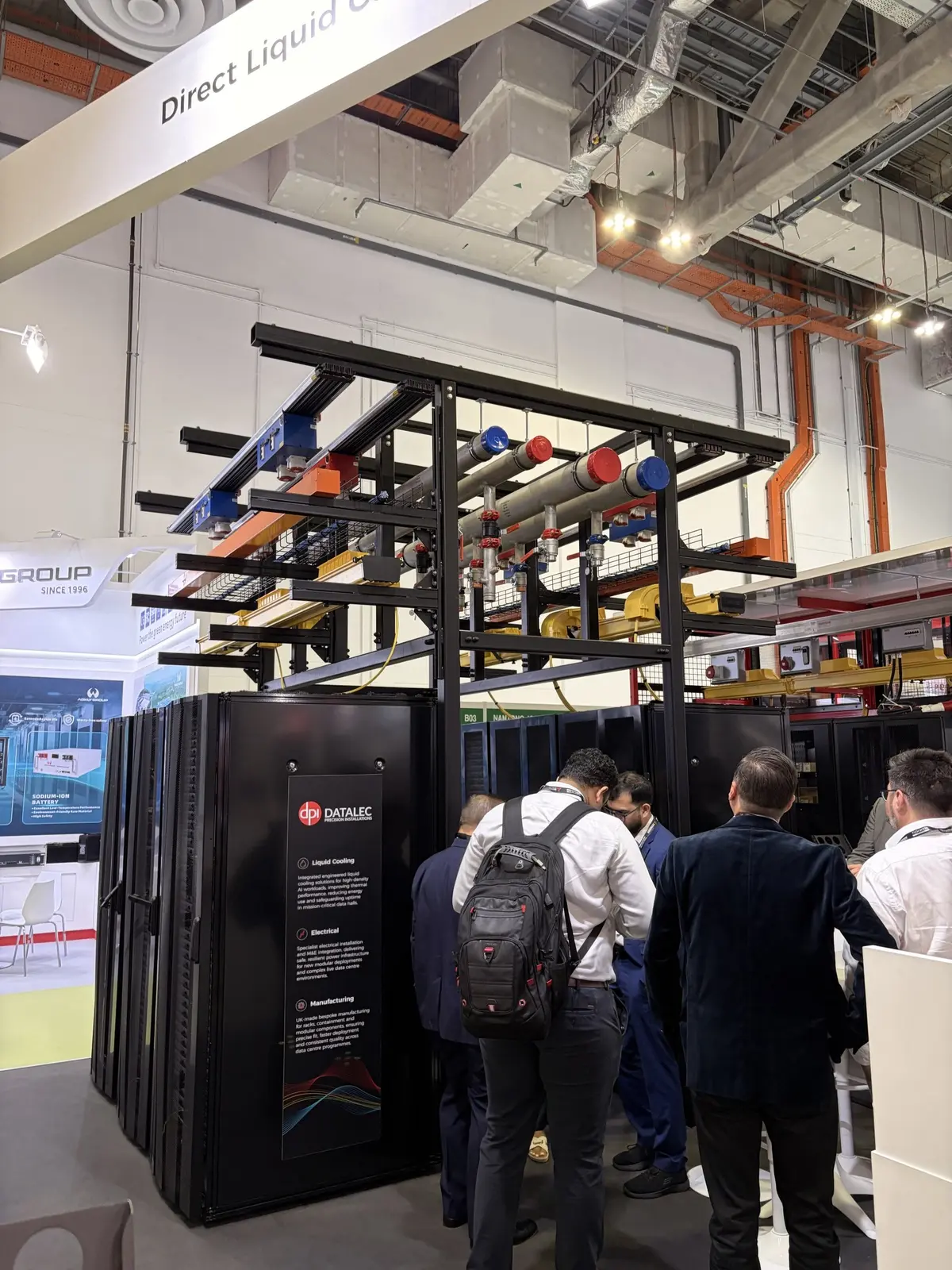 Visitors at the Datalec booth under liquid-cooling pipework running above a row of racks.